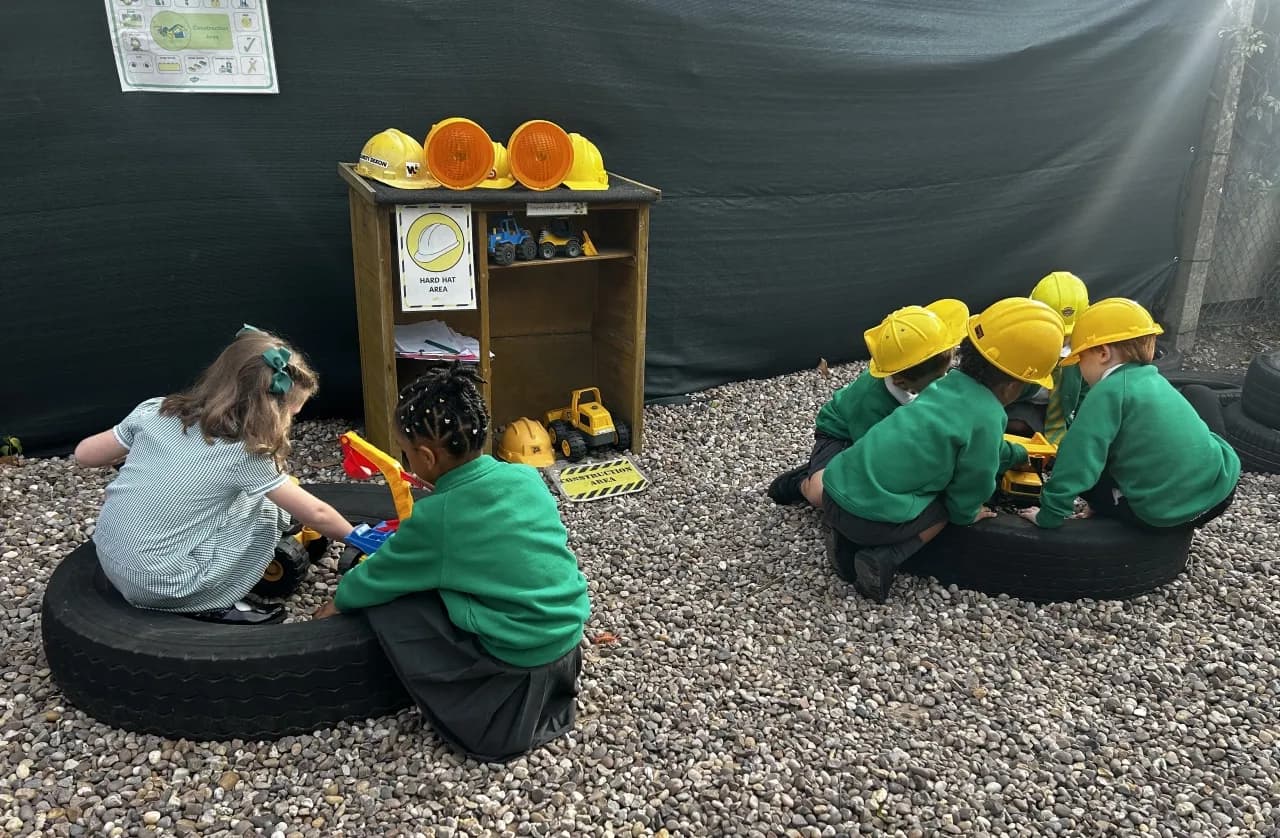 Several young children in green shirts and some in yellow construction hats are playing with toy construction vehicles on a gravel ground, with a wooden shelf holding more toys and hard hats in the background.